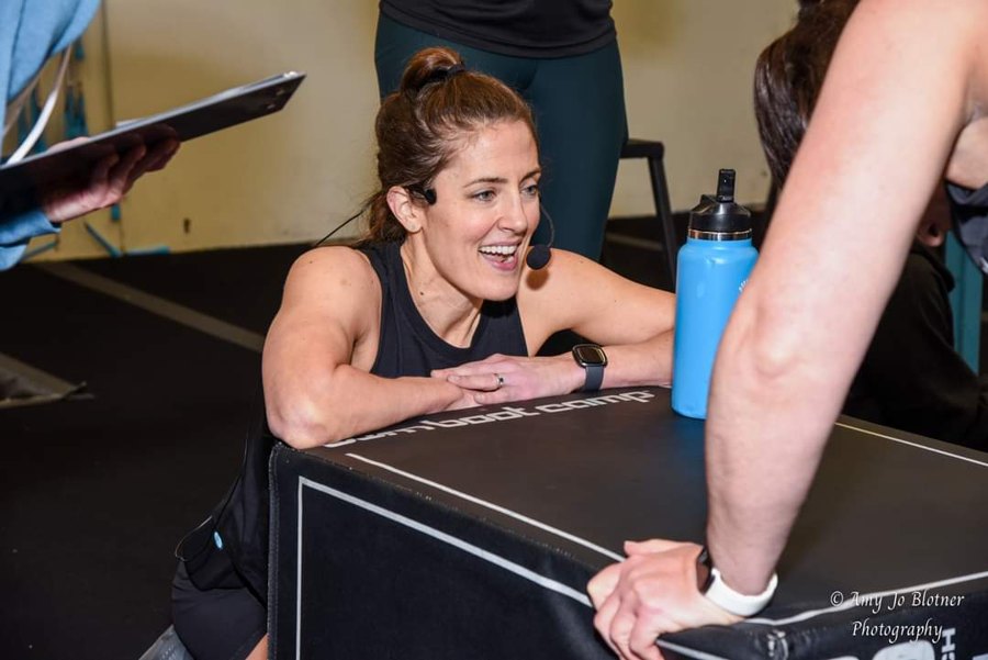 Caitlin mid-session, coaching headset on, leaning on a plyo box and laughing with a client
